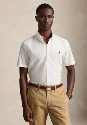 Mężczyzna w białej koszuli z krótkim rękawem z małym logo, beżowych spodniach, brązowym pasku i zegarku, stojący z jedną ręką w kieszeni.