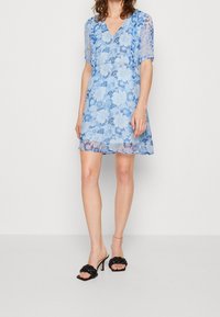 Robe florale bleue avec des manches courtes transparentes et un décolleté en V. Fabriquée en dentelle, elle possède un corsage ajusté et une jupe fluide, accompagnée de sandales à talons noires.