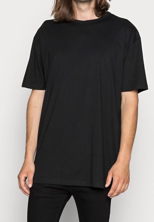 Homme aux cheveux mi-longs, portant un t-shirt noir uni oversized et un pantalon noir, debout devant un fond blanc uni.