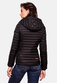 Veste matelassée noire avec capuche, présentant une finition texturée, une coupe ajustée, et un accent rouge près de la poche ; étiquette brune sur l'ourlet.