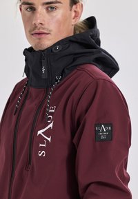 Bordoux jack met een zwarte capuchon, voorzien van een wit logo aan de voorkant en een label op de mouw. Glad materiaal met verstelbare trekkoorden.