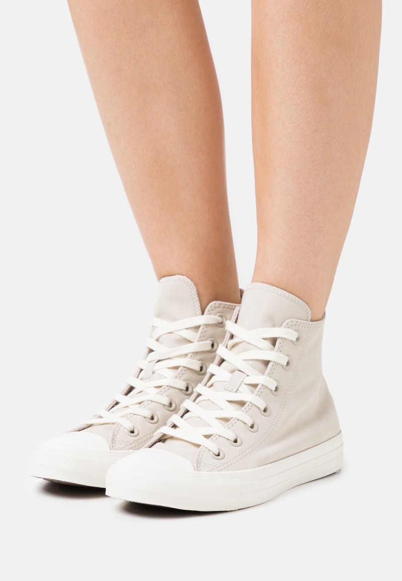 Vysoké tenisky z lehkého béžového plátna s bílými tkaničkami a gumovými podrážkami. Mají kulatou špičku a švy na límci.