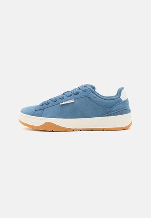 Niski niebieski zamszowy sneaker z białą podeszwą, gumową podeszwą zewnętrzną i sznurowanym zapięciem, z małą tkaną metką "Tommy Jeans" po boku.