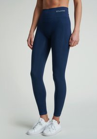 Granatowe legginsy z wysokim stanem, o prążkowanej fakturze i elastycznym pasie, w połączeniu z białymi sportowymi butami. Prosty design, bez wzorów.