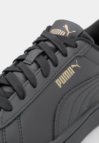 Mörkgrå lädersneaker med svarta snörningar, guldig "PUMA"-logga och en texturerad sidopanel. Rena linjer och enkel design.