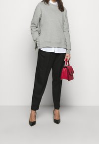 Grauer Pullover mit geripptem Saum und seitlichen Rüschen, über einem weißen Hemd getragen. Schwarze Hose, schwarze Absatzschuhe und eine rote Handtasche runden das Outfit ab.
