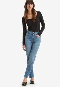 Body preto de mangas longas emparelhado com jeans de denim azul de cintura alta, apresentando um acabamento desgastado e tachas decorativas nos bolsos. Sapatos pretos.