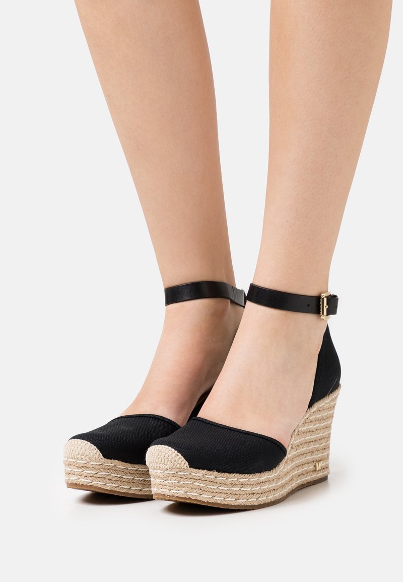 Jambes portant des espadrilles compensées en tissu noir avec des brides à la cheville et des semelles en corde tressée beige sur fond blanc.