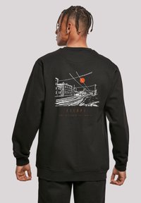 Person mit geflochtenen Haaren, der wegschaut, trägt einen schwarzen Pullover mit weißer Linienzeichnung von Bahngleisen, Gebäuden, Oberleitungen und einer orangefarbenen Sonne auf dem Rücken.