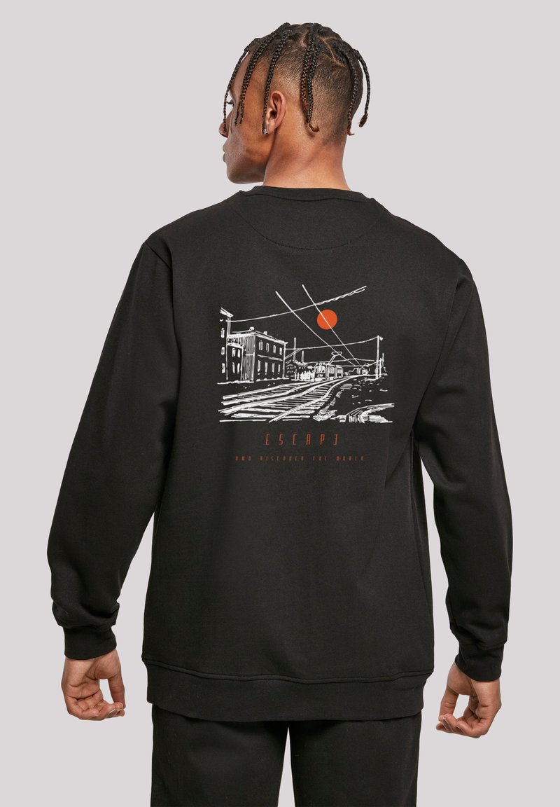 Person mit geflochtenen Haaren, der wegschaut, trägt einen schwarzen Pullover mit weißer Linienzeichnung von Bahngleisen, Gebäuden, Oberleitungen und einer orangefarbenen Sonne auf dem Rücken.