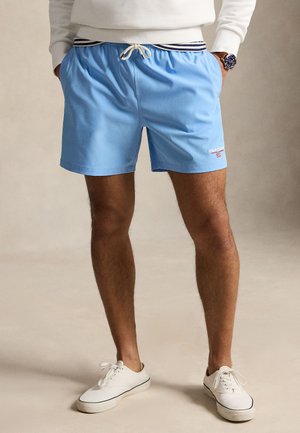 Hombre con pantalones cortos azul claro con cinturilla a rayas y zapatillas blancas, con las manos en los bolsillos, de pie sobre un fondo neutro.