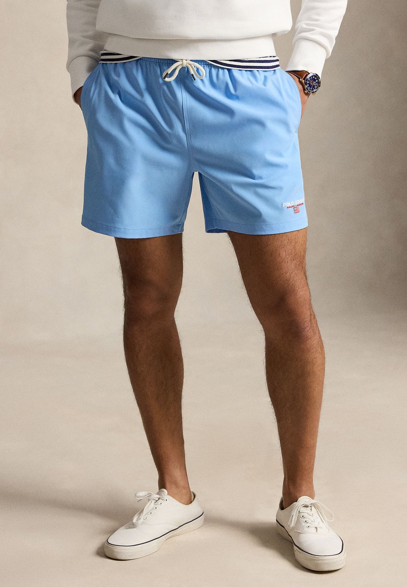 Mees, kes kannab helesiniseid lühikesi pükse triibulise vöökohaga ja valgeid tossusid, käed taskus, seisab neutraalsel taustal.