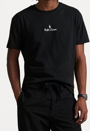 Mann trägt ein schwarzes Ralph Lauren T-Shirt mit weißem Logo, schwarze Hose mit gebundenem Gürtel und eine schwarze Armbanduhr, steht mit den Händen in der Nähe der Taschen.