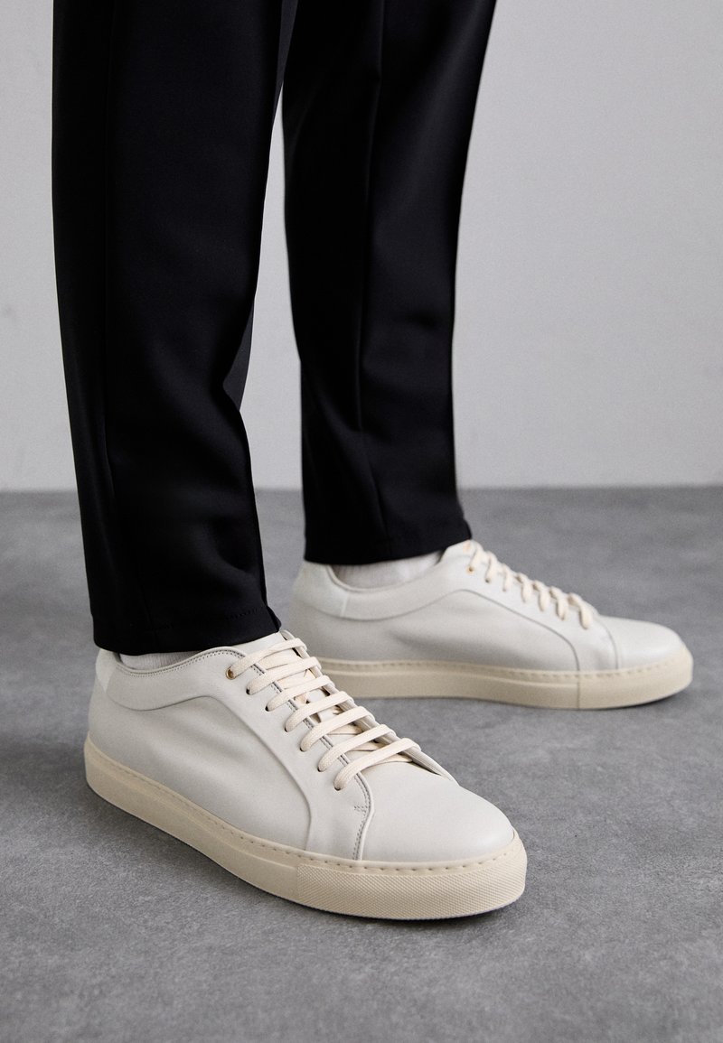 Zapatillas de cuero color beige claro con cordones planos, una superficie texturizada y una suela de goma color crema en contraste, combinadas con pantalones negros ajustados.