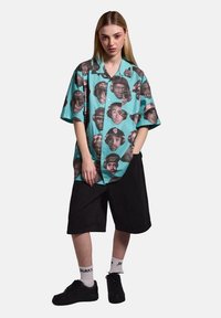 Short-sleeve button-up shirt in turquoise, featuring a repeated black-and-white face print. Paired with black shorts and black shoes.