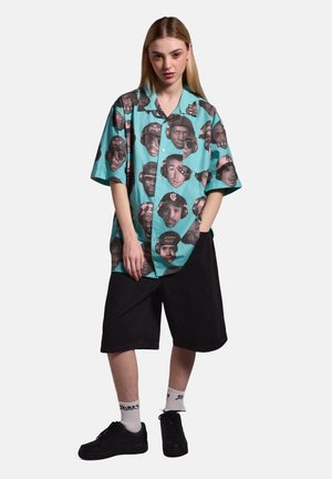 Kurzärmliges Button-up-Hemd in Türkis, mit einem wiederholten Schwarz-Weiß-Gesichtsdruck. Kombiniert mit schwarzen Shorts und schwarzen Schuhen.