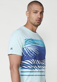 Ljusblå bomullströja med en blå och vit palmblads grafik över bröstet och en logotyp på vänster ärm. Korta ärmar.