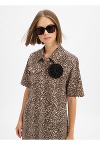 Beige shirtjurk met luipaardpatroon en korte mouwen, voorzien van een zwart stoffen bloemenaccent op de borst en een knoopsluiting.