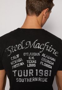 Czarny bawełniany t-shirt z wyblakłym szarym wzorem graficznym, na którym znajduje się napis "Steel Machine TOUR 1981" oraz różne stany USA w stylu zniszczonym.
