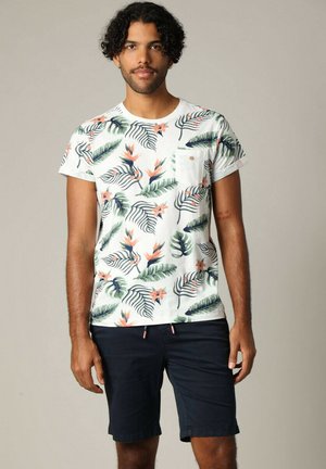 Chemise blanche à manches courtes avec un motif de feuilles tropicales et de fleurs, dotée d'une poche poitrine et d'un subtil détail en métal doré. Assortie avec un short foncé.