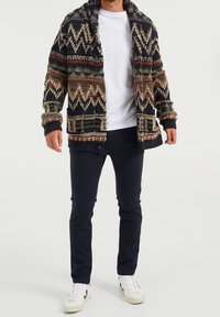 Hombre vestido con un cárdigan de punto con patrón en azul marino, camiseta blanca, pantalones oscuros ajustados y zapatillas blancas, de pie contra un fondo blanco.