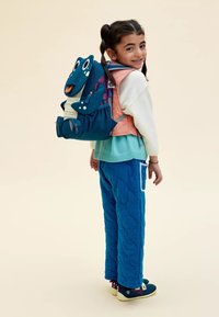 Zaino per bambini blu con design di rana, accenti colorati e diversi scomparti con cerniera. Bambino vestito con pantaloni trapuntati blu e maglione.