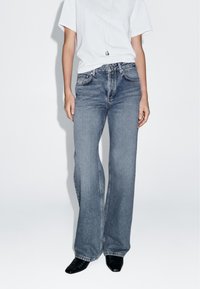 Hochtaillierte Weitbeine-Jeans aus mittlerem blauen Denim mit dezenten Ausbleichungen. Kombiniert mit einem schlichten weißen T-Shirt und schwarzen High Heels.