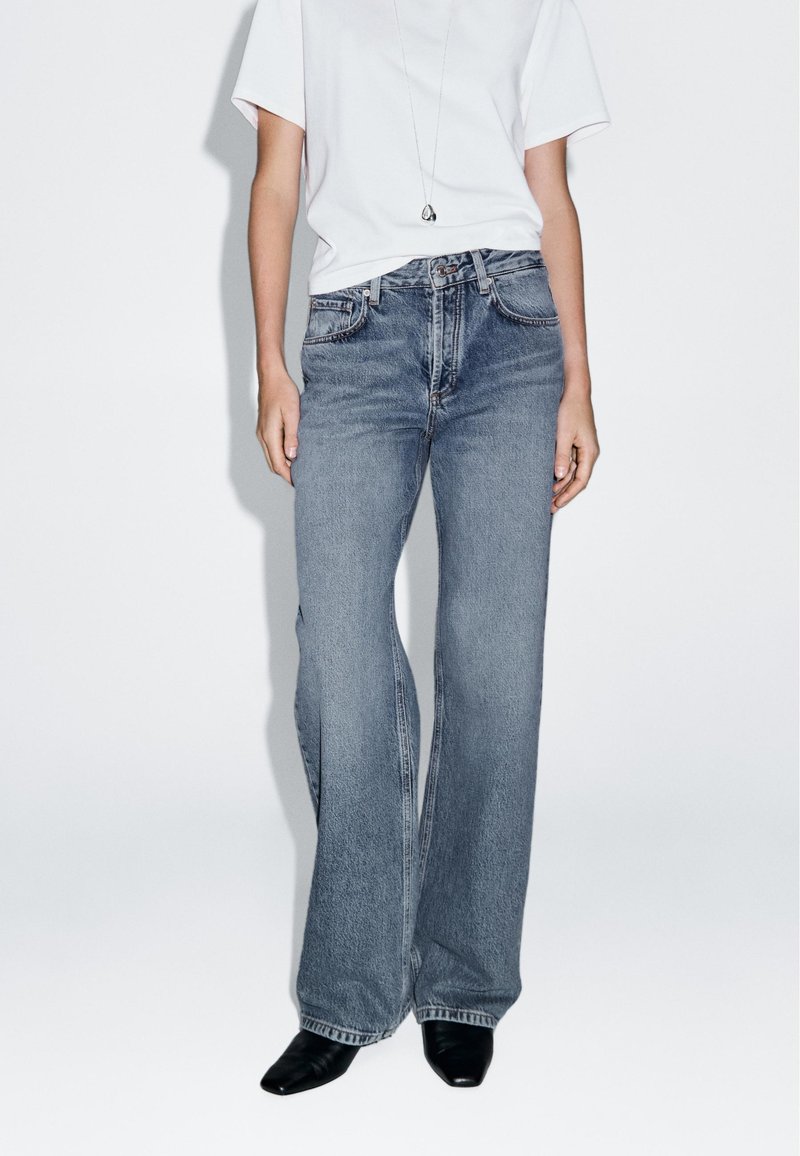 Hochtaillierte Weitbeine-Jeans aus mittlerem blauen Denim mit dezenten Ausbleichungen. Kombiniert mit einem schlichten weißen T-Shirt und schwarzen High Heels.