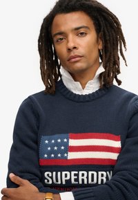 Maglione a maglia blu navy con un motivo testurizzato della bandiera americana in rosso, bianco e blu, con la scritta "SUPERDRY" in bold bianco.