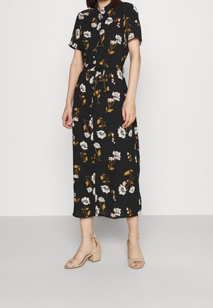 Vrouw in een zwarte midi-jurk met witte en bruine bloemenprint, korte mouwen, knoopsluiting aan de voorkant en beige sandalen met blokhak.