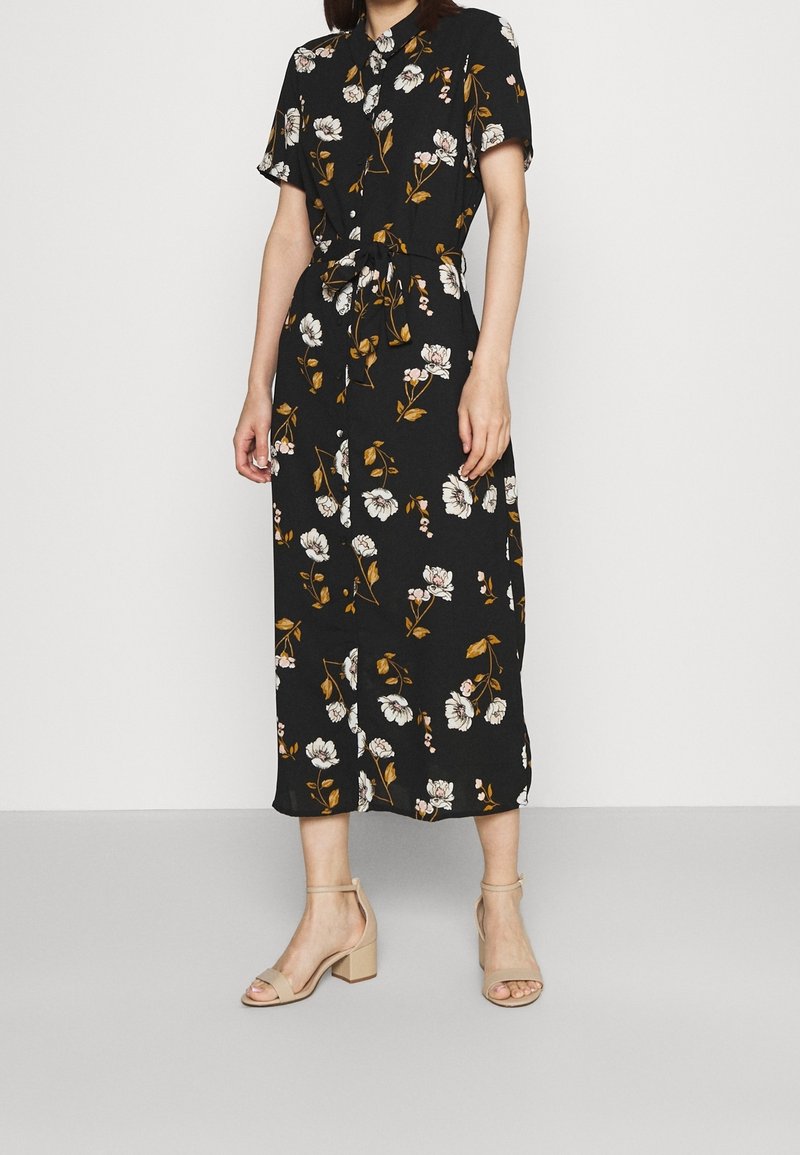 Femme portant une robe midi noire à imprimé floral blanc et marron, manches courtes, boutonnée à l'avant, et des sandales à talon bloc beige.