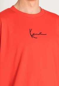 Červené bavlněné tričko s kulatým výstřihem, na hrudi s černým vyšitým podpisem. Hladká textura, krátké rukávy.