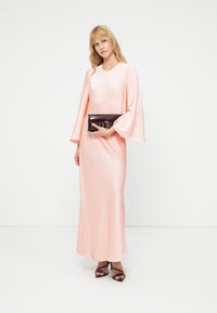 Μακρύ ροζ παστέλ σατέν φόρεμα με μανίκια καμπάνα, στρογγυλή λαιμόκοψη και μήκος μέχρι το πάτωμα, συνδυασμένο με σκούρο τσαντάκι χειρός και μαύρα τακούνια.