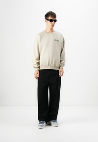 Sweatshirt gris avec des poignets côtelés, associé à un pantalon noir ample et des baskets bleues. Le modèle porte des lunettes de soleil noires pour un look décontracté.