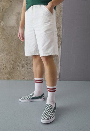 Valkoiset puuvillahousut sivutaskuilla, yhdistettynä valkoisiin sukkiin, joissa on punaiset raidat, ja vihreävalko-ruudullisiin slip-on-kenkiin.