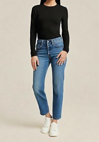 Femme portant un haut noir ajusté à manches longues, un jean bleu taille haute et des baskets blanches à lacets, debout devant un fond uni.