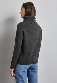 Grå stickad tröja med hög, ribbad krage. Mjuk textur, avslappnad passform och långa ärmar. I kombination med ljusblå jeans. Bakifrån.