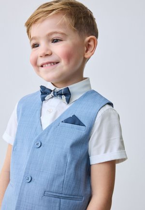 Lachende jonge jongen met een lichtblauw vest, wit overhemd met opgerolde mouwen, en een marineblauw gebloemd strikje en pochet.