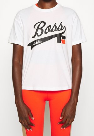 Osoba o ciemnej karnacji, ubrana w biały t-shirt "Boss Athletic" oraz jaskrawo czerwone legginsy sportowe, stojąca z rękami swobodnie opuszczonymi wzdłuż ciała.