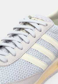 Gros plan sur une basket en tricot bleu clair avec des empiècements en daim beige, trois bandes crème et des lacets blancs.