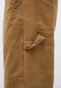 Καφέ παντελόνι από καμβά με πλαϊκή τσέπη, ορατές ραφές και μια μικρή λεπτομέρεια σε θηλιά. Ο υφασμένος καμβάς έχει ελαφρώς φθαρμένη εμφάνιση.