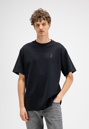 Jeune homme aux cheveux bouclés, portant un t-shirt noir et un jean gris, debout devant un fond blanc uni.
