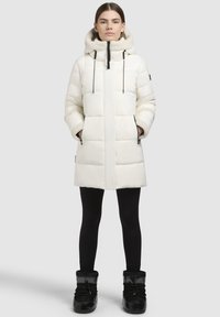 Vit vadderad jacka med mjuk, pälsfodrad huva, framsida med dragkedja och toggeldragningar. Buren över svarta leggings och glänsande svarta stövlar.