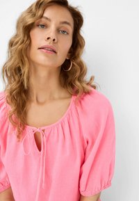 Blusa rosa a maniche corte con scollatura rotonda, maniche elastiche e dettaglio a fiocco nella parte anteriore. Il materiale sembra leggero e testurizzato.