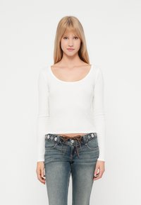 Νέα γυναίκα με ίσια ξανθά μαλλιά φοράει ένα λευκό μπλουζάκι με μακριά μανίκια και στρογγυλή λαιμόκοψη, καθώς κ�αι μπλε τζιν με ζώνη με λουλουδάτα κεντήματα.