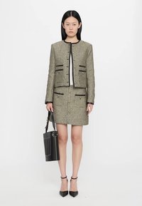 Tweed jacket and skirt set in olive with black trim; features gold buttons, pockets, and textured fabric. Model holds a black handbag.