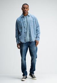Light blue corduroy shirt jacket with two chest pockets, layered over a gray t-shirt; paired with straight-leg blue jeans and black sneakers.