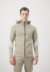 Giacca beige con cappuccio e dettagli grigi, con zip frontale, tasche laterali e logo Puma. Realizzata in tessuto morbido e testurizzato per il comfort.