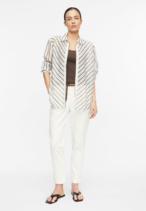 Woman standing wearing a white striped button-up shirt over a brown top, white pants, brown woven belt, and black open-toe sandals.