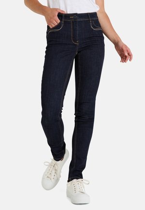 Jean en denim bleu foncé avec une coupe slim, présentant des coutures orange, des poches avant et une fermeture à bouton. Portés avec des baskets blanches.